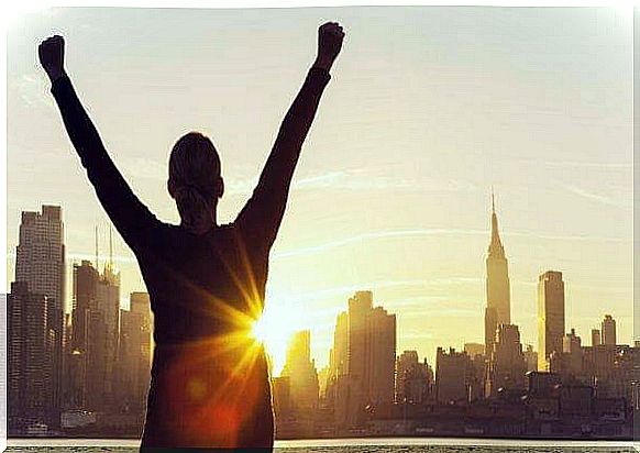 A woman gazes at a city skyline at sunset and holds her arms in the air. 